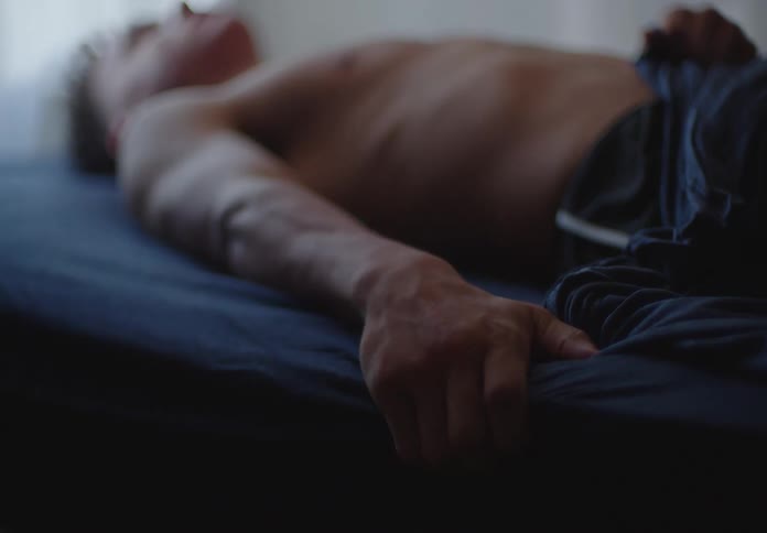 A shallow focus shot of a man's arm and hand as he lies shirtless on a bed in a dimly lit room