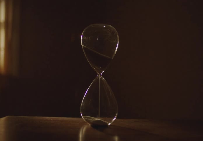 A slow pull-back shot reveals an hourglass measuring the passing of time on a wooden table in a dark, moody room