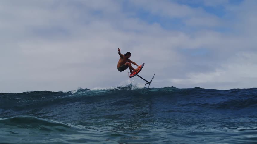 A hydrofoil surfer gracefully launches off a deep blue ocean wave in stunning super slow motion
