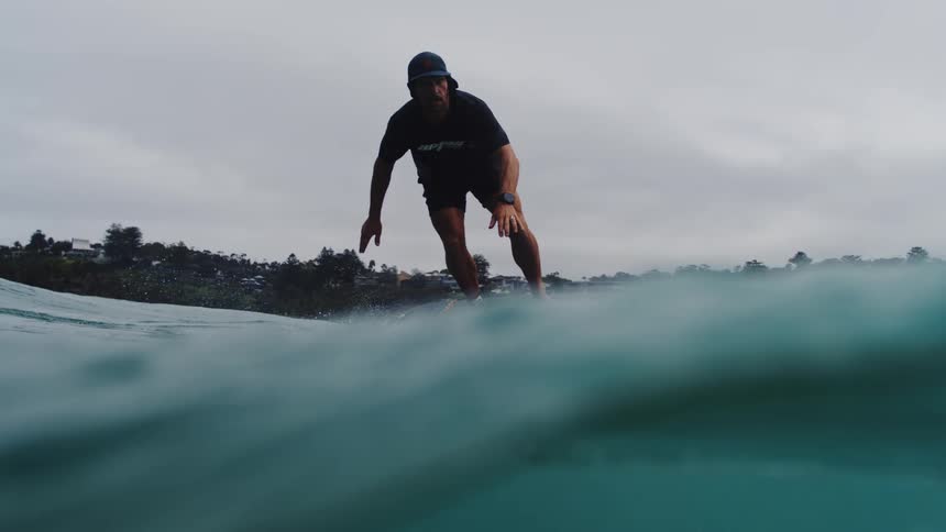 A man skillfully rides an electric hydrofoil surfboard in slow motion across the ocean towards a sandy beach