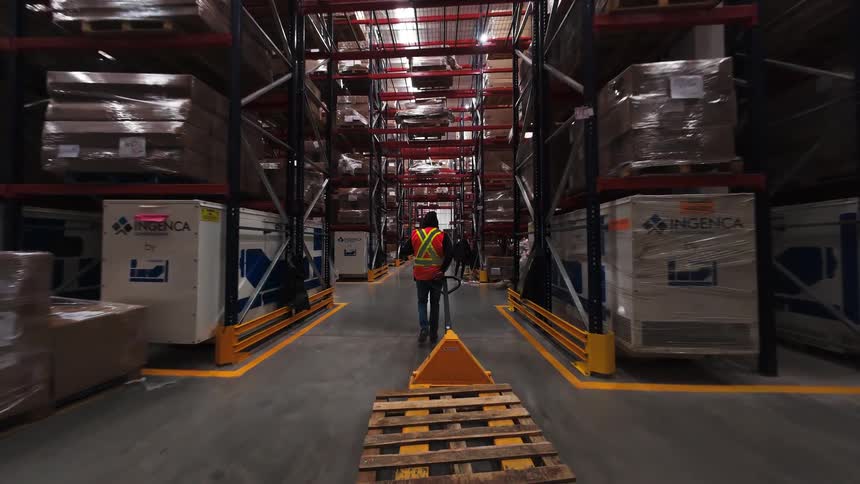 A worker in a safety vest pulls a manual pallet jack through the long aisles of a vast distribution warehouse