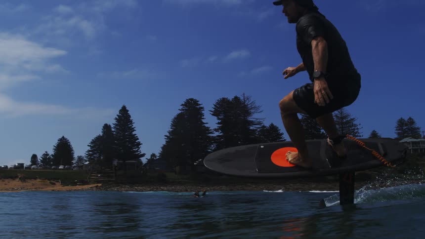 A man gracefully rides an electric hydrofoil surfboard in slow motion over the ocean, with the coastline visible under a bright blue sky