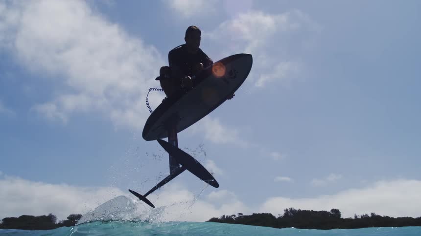 A man on a hydrofoil surfboard soars out of the water in a dramatic slow-motion silhouette against the bright sky before splashing back down