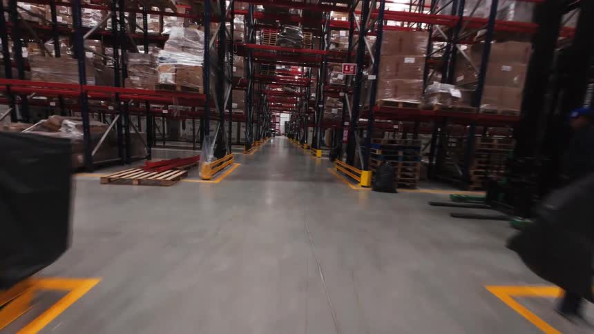 A dynamic drone shot flies through the aisles and between the racks of a large, modern logistics warehouse filled with stacked inventory