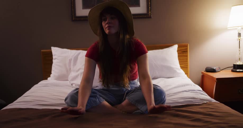 A young woman wearing a straw hat sits contemplatively on a bed in a dimly lit motel room