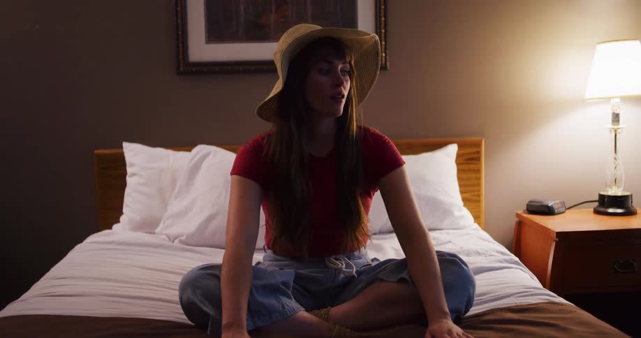 A playful young woman wearing a sun hat sits cross-legged on a hotel bed before standing up to jump