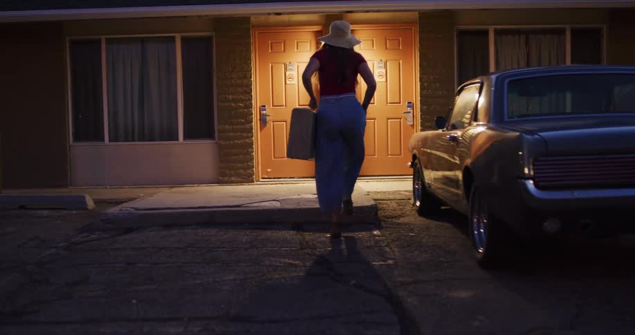 A woman in a stylish hat takes her vintage suitcase from the trunk of a classic car and walks into her motel room at night