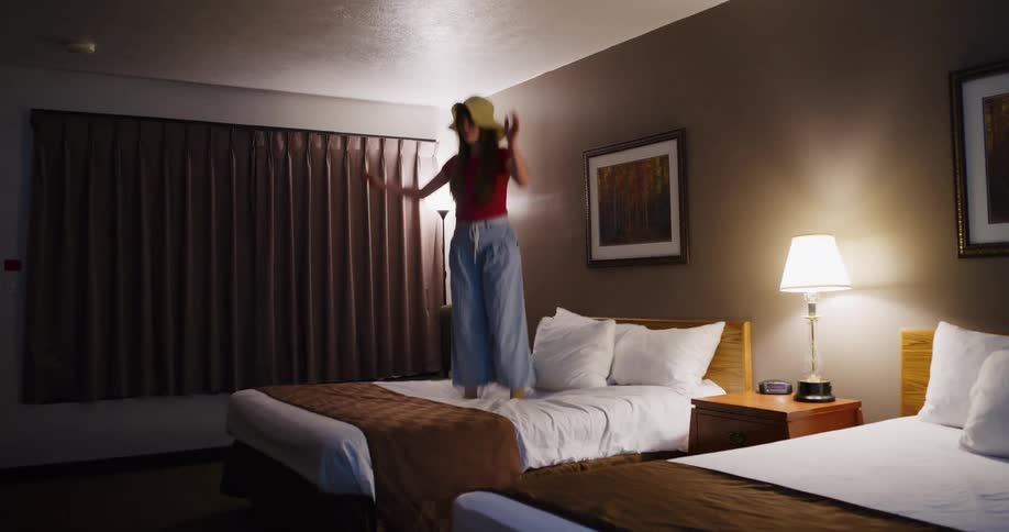 A carefree young woman in a sun hat joyfully dances and jumps on the bed in her motel room in slow motion