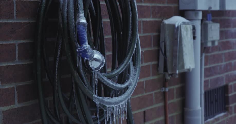 A frozen garden hose with icicles hangs against a brick wall as snow falls gently