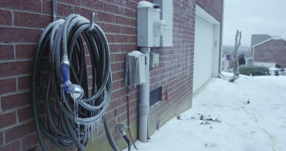 A frozen green garden hose with icicles hanging from the nozzle is mounted on a brick wall during a cold winter day