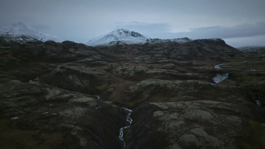 A slow aerial push-in over a vast, mossy tundra with a winding river leading towards snow-capped mountains on a gloomy day