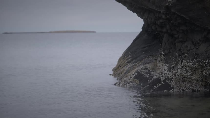 A serene and minimalist view of a calm sea lapping against a large rock, with a distant island on the horizon under an overcast sky