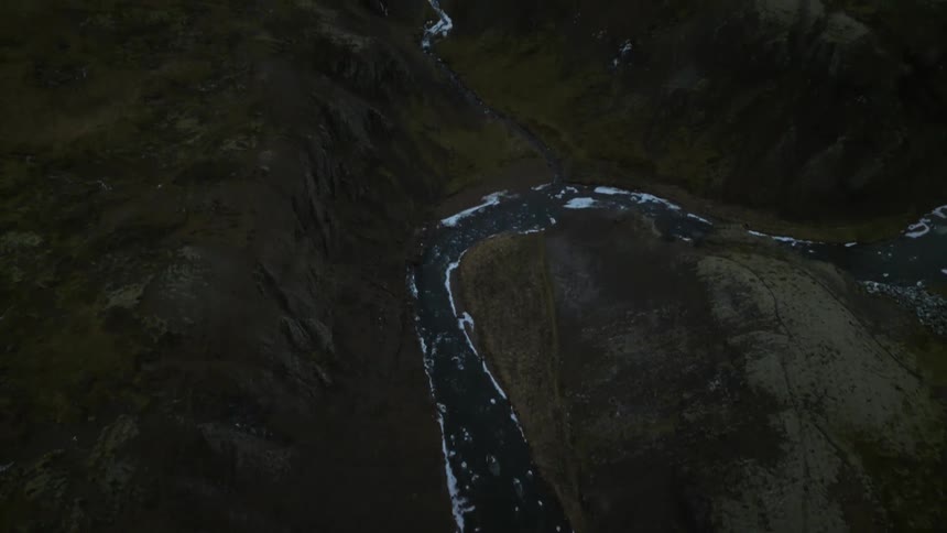A dramatic aerial view reveals a partially frozen river winding through a dark, mossy landscape toward a distant snow-capped mountain