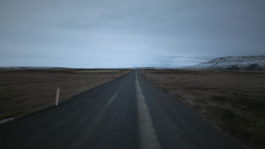 A point-of-view shot driving down a long, straight gravel road through a barren, wintery landscape in Iceland under an overcast sky