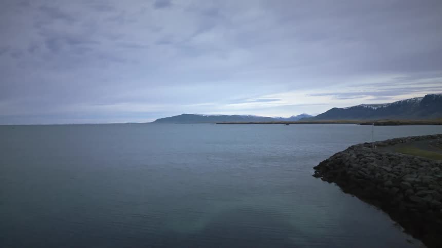 Slow aerial dolly shot over the calm ocean waters of an Icelandic harbor on an overcast day with snow-capped mountains in the distance