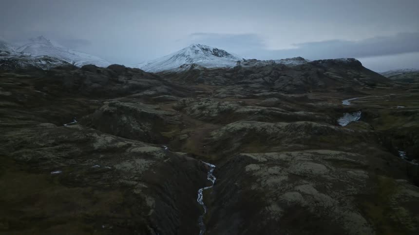 A slow aerial pullback reveals a vast and desolate Icelandic landscape with a winding river and a snow-capped mountain on the horizon
