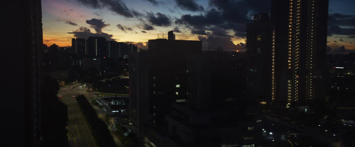 A dramatic sunset glows behind a silhouetted city skyline with illuminated buildings and roads at dusk