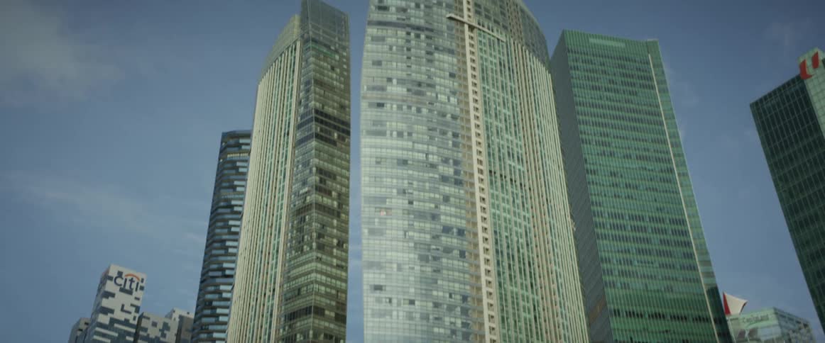 A slow tilt-up reveals the towering glass facades of modern skyscrapers against a clear blue sky, rising from behind a line of lush green trees