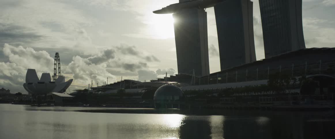 A slow tilt-up shot reveals the iconic Marina Bay Sands hotel in Singapore, silhouetted against the bright morning sun reflecting off the calm water
