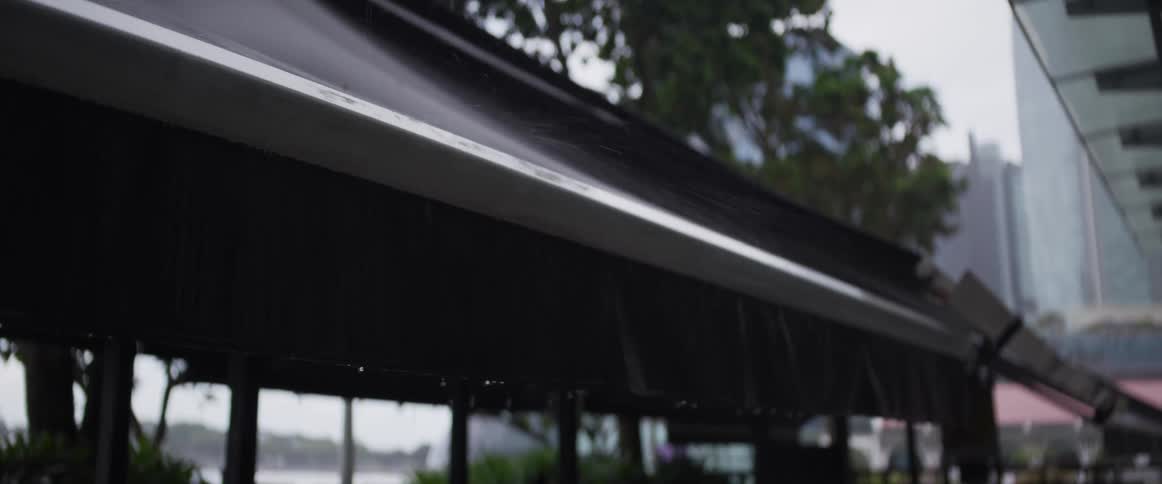 Heavy rain falls onto a black outdoor awning, splashing and dripping off the edge on a gloomy day