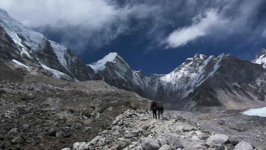 White fluffy clouds and a blue sky over a snow capped mountain range as people walk peacefully