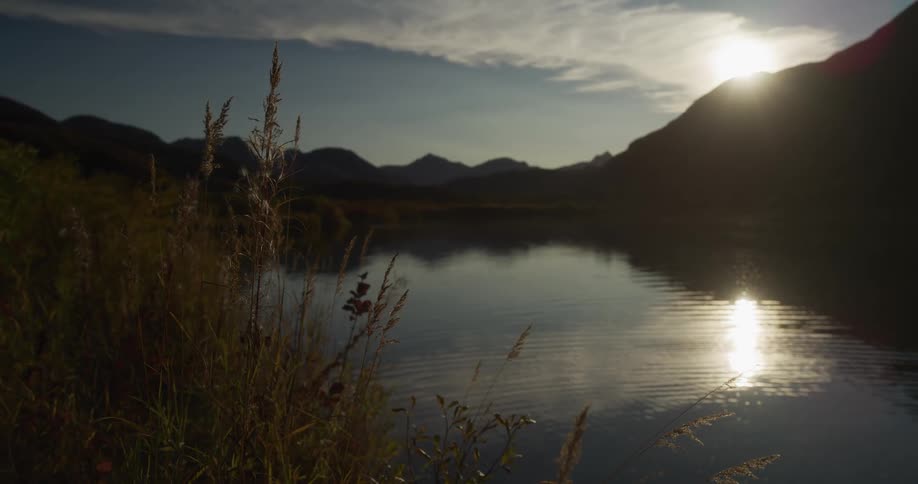 A tranquil lake reflects the setting sun and silhouetted mountains from a grassy shore