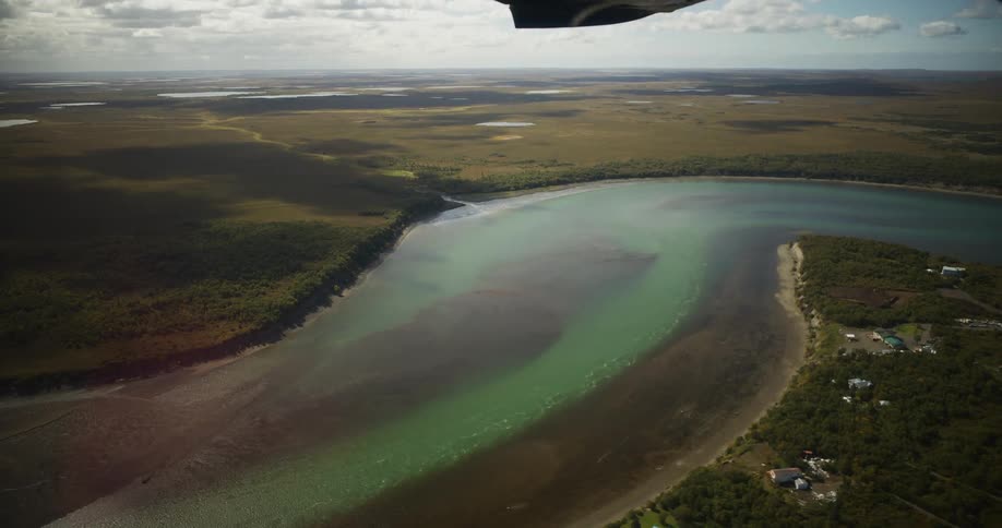 An aerial view from an airplane reveals a vast, remote landscape where a turquoise river meets the coast next to a small settlement
