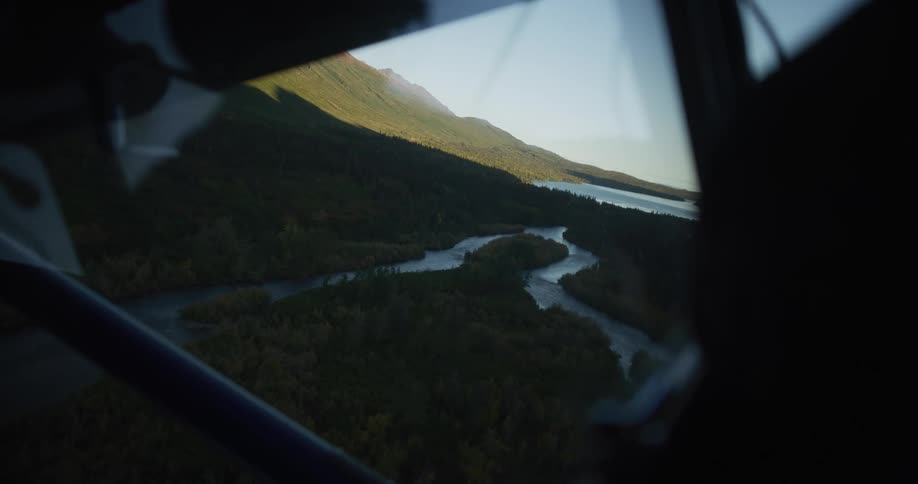Aerial view from a small plane flying over a lush green wilderness with a winding river and mountains at sunset