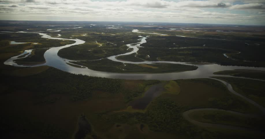 An aerial view captures a vast and remote landscape where a wide, meandering river snakes through forests and tundra under a partly cloudy sky