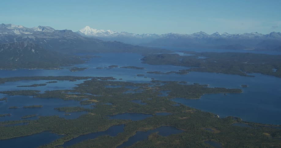 A stunning, static aerial view of a vast wilderness landscape featuring numerous interconnected lakes, forested islands, and a distant snow-capped mountain range under a clear blue sky