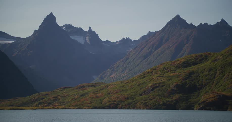 A slow, majestic pan reveals a stunning Alaskan mountain range with jagged peaks rising above a tranquil lake and rolling, autumn-colored hills