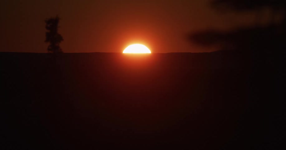 A timelapse of a brilliant orange sun setting behind a silhouetted, hazy mountain horizon until the landscape is completely dark
