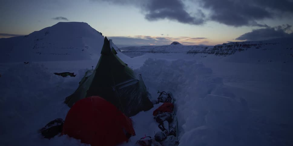 A remote campsite with tents is nestled in a vast, snowy mountain landscape during twilight