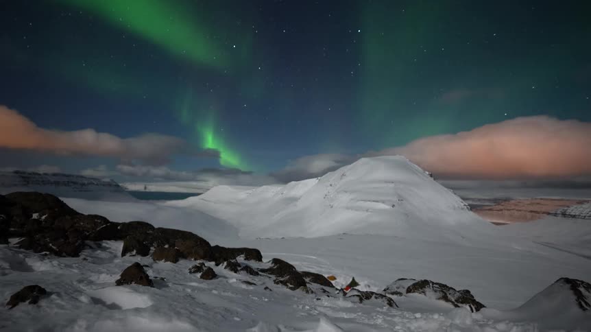 A breathtaking timelapse captures the vibrant green Northern Lights dancing across the starry night sky above a vast, snow-covered mountain landscape