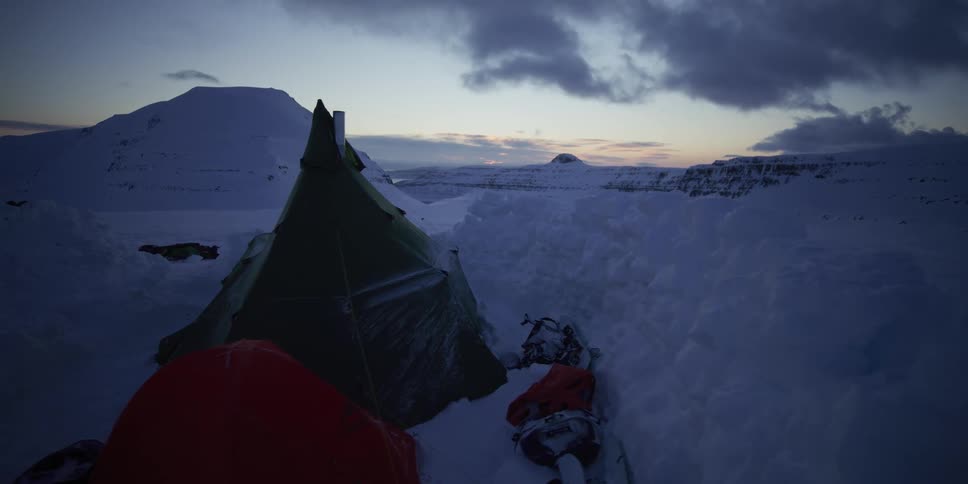 Dolly shot reveals a quiet and cold winter campsite with tents pitched in deep snow amongst the mountains at dusk