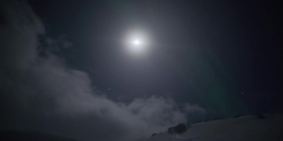 A majestic time-lapse of the moon and a faint aurora borealis as clouds drift over a snowy mountain landscape at night