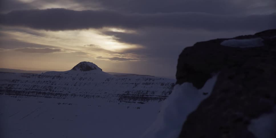 A vast snowy mountain landscape seen from a cliffside under a dark, moody sky at sunset