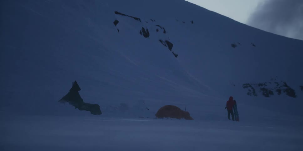 A snowboarder stands at a remote campsite on a snowy mountain as the wind blows snow during the cold, dark evening