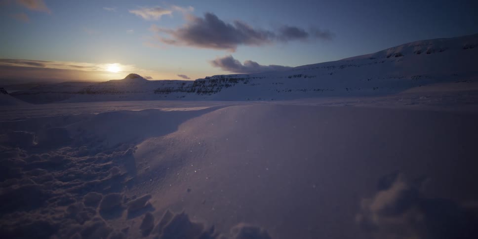 A slow dolly shot glides over a vast, snow-covered landscape with mountains in the distance as the sun sets on the horizon