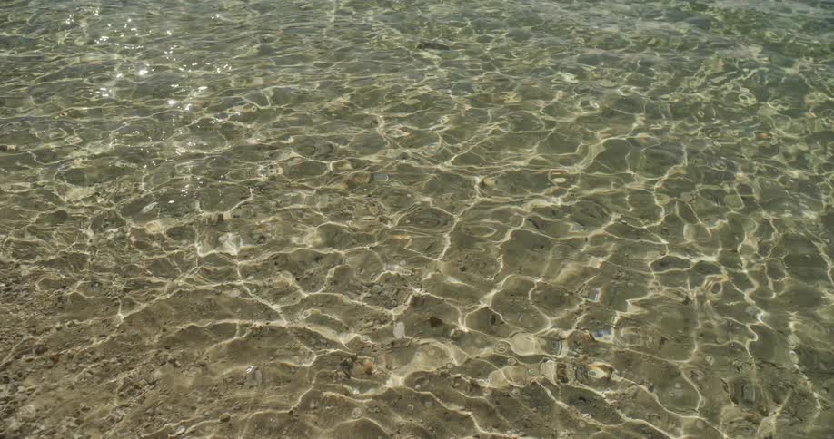 Calm, clear water ripples gently in the sun, creating sparkling light reflections on the sandy sea floor
