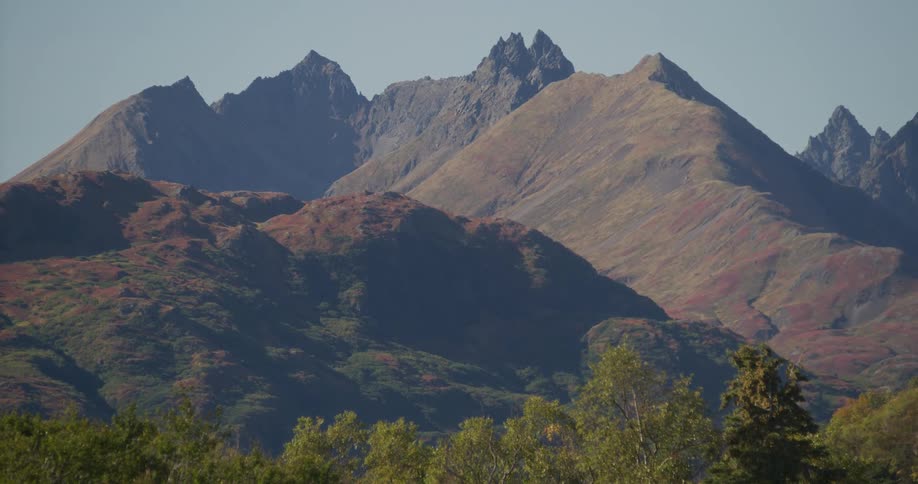 A static landscape shot shows a majestic mountain range with rugged peaks and colorful autumn foliage on a clear, sunny day