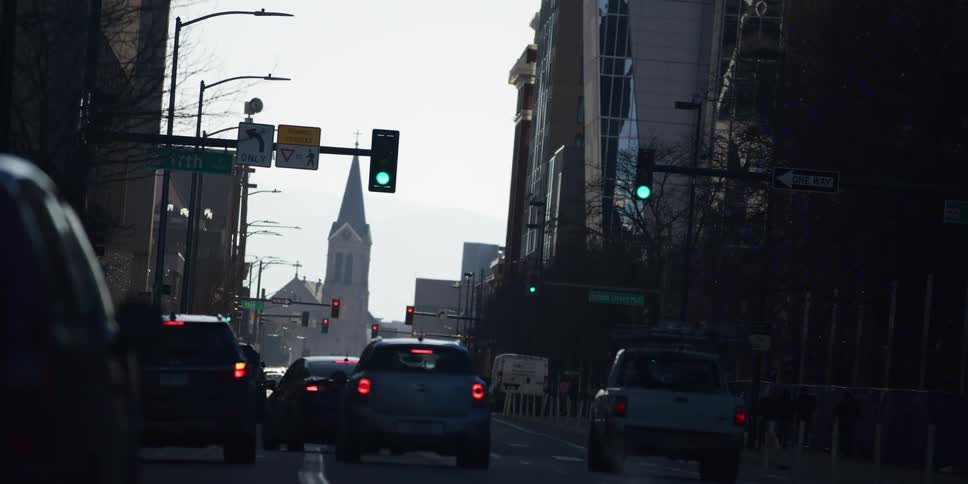 Backlit city traffic waits at a stoplight with a church steeple in the distance as the light changes from green to yellow