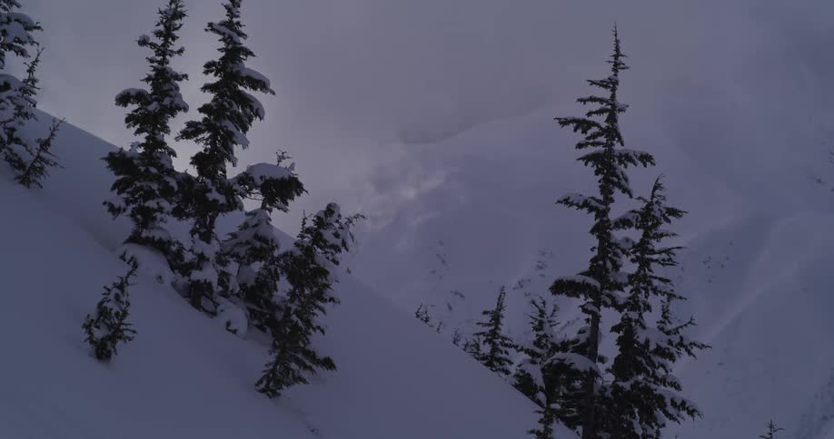 Dark, ominous clouds move swiftly over a snowy mountain landscape dotted with evergreen trees