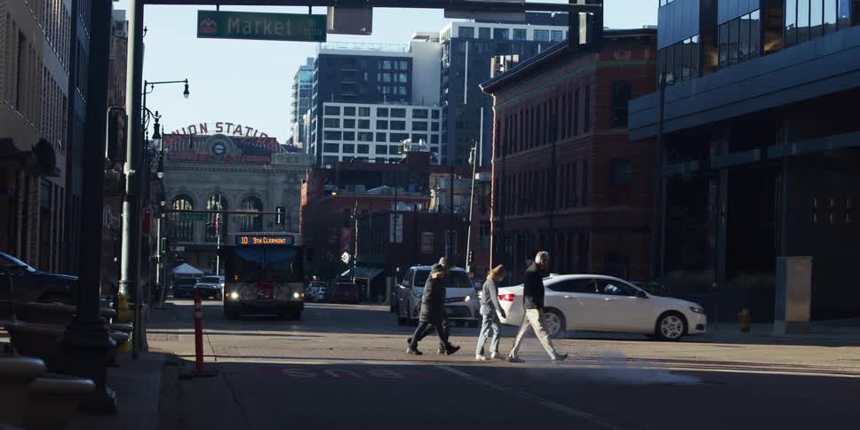 Pedestrians cross a sunlit city street in slow motion with traffic and Denver's Union Station in the background