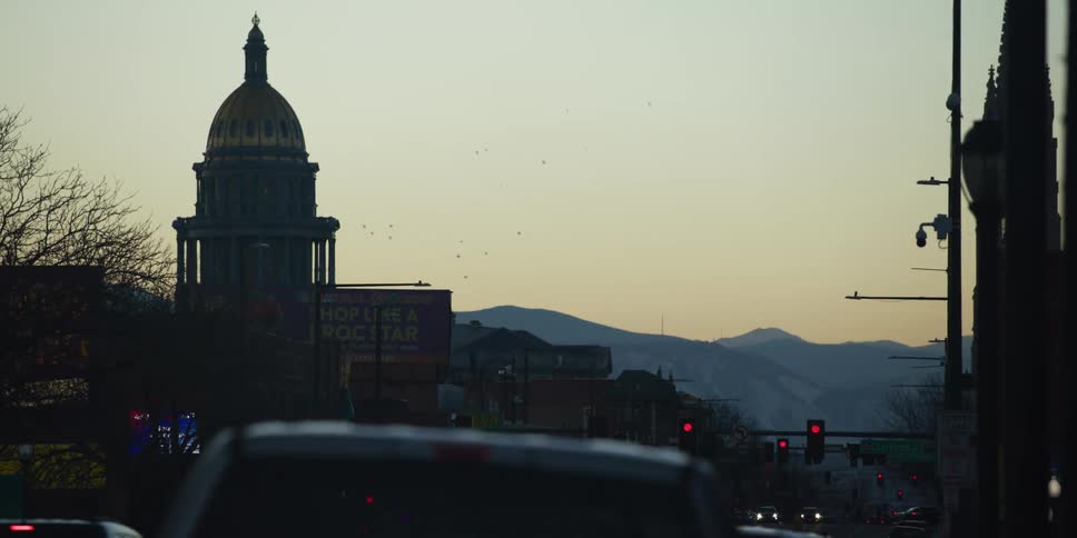 The Colorado State Capitol building is silhouetted against the Rocky Mountains at dusk as traffic navigates the city streets