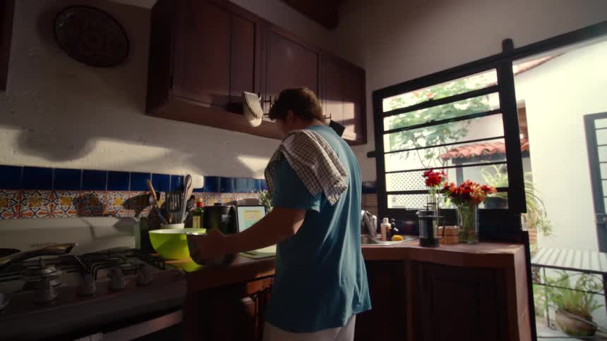 A man with a dish towel over his shoulder walks into a sunlit rustic kitchen and opens a cabinet to begin preparing a meal