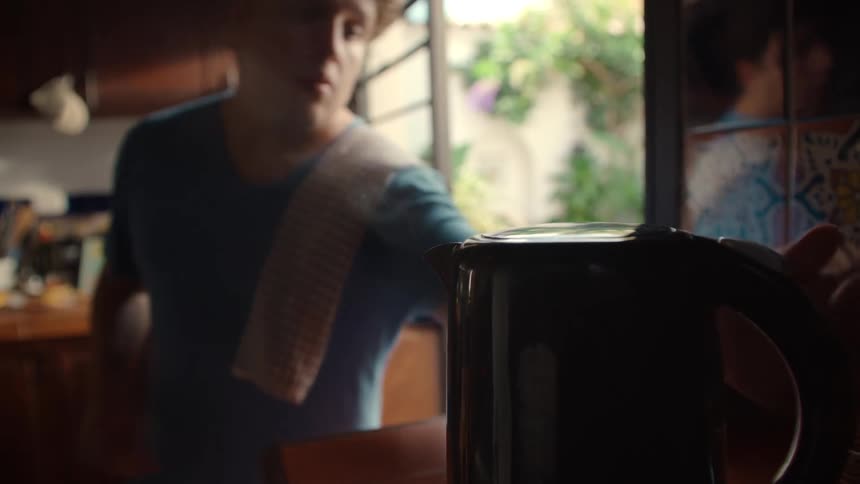 A man grinds coffee beans and then pours steaming hot water from a kettle into a coffee press in a sunny kitchen