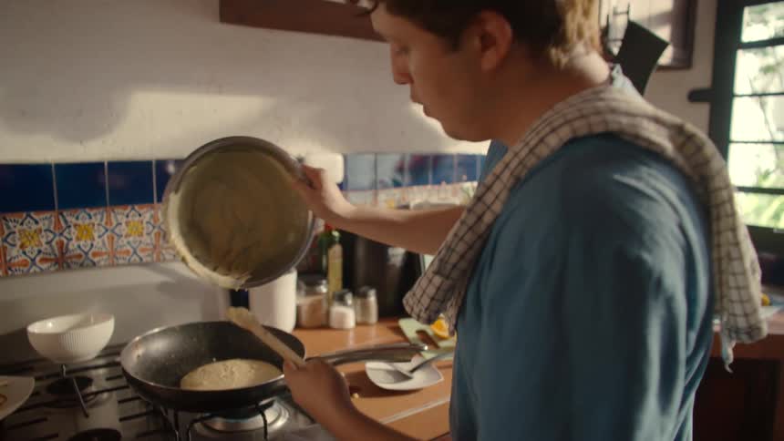 Slow motion shot of a man carefully pouring pancake batter from a large bowl into a hot frying pan on a gas stove