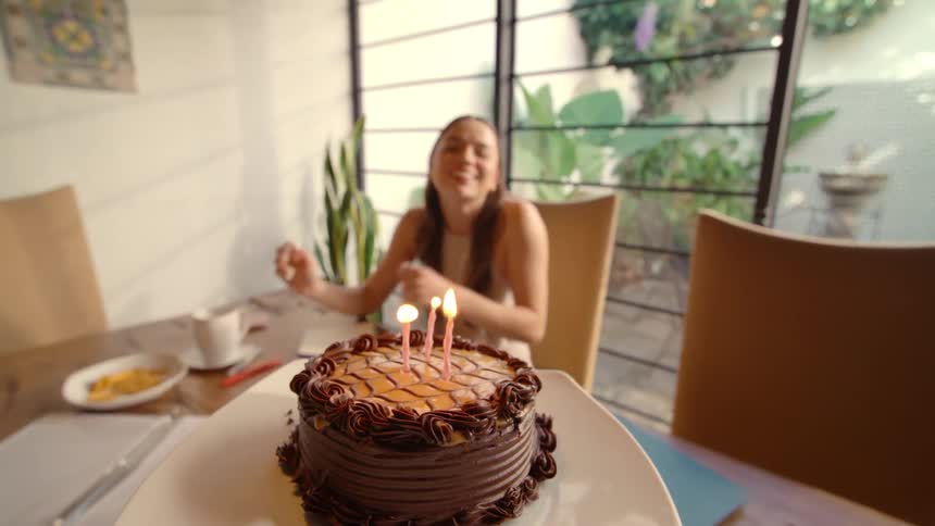 From a first-person perspective, a birthday cake with lit candles is brought to a smiling woman who joyfully receives the surprise