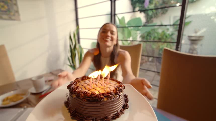 A first-person view of bringing a chocolate birthday cake with lit candles to a surprised and happy woman at a dining table